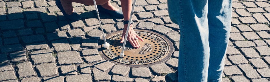 Eine blinde Frau ertastet den Gullideckel des "Bremer Lochs". Wirft man Geld in den Einwurfschlitz, ertönen Stimmen der Bremer Stadtmusikanten. 