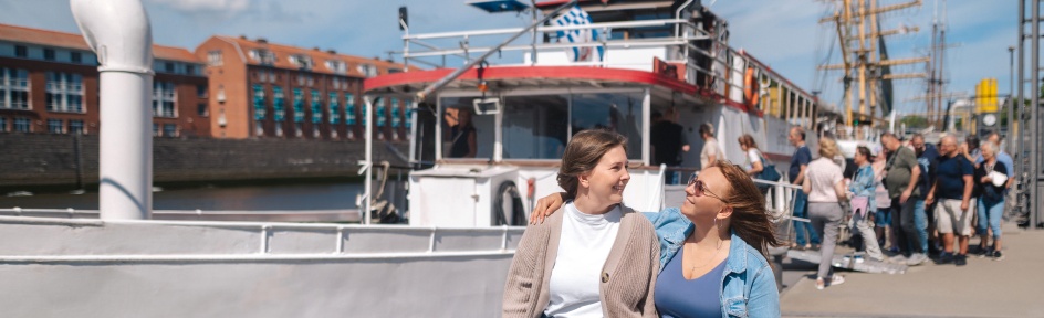 Two women laugh at each other. In the background, people are boarding a ship.