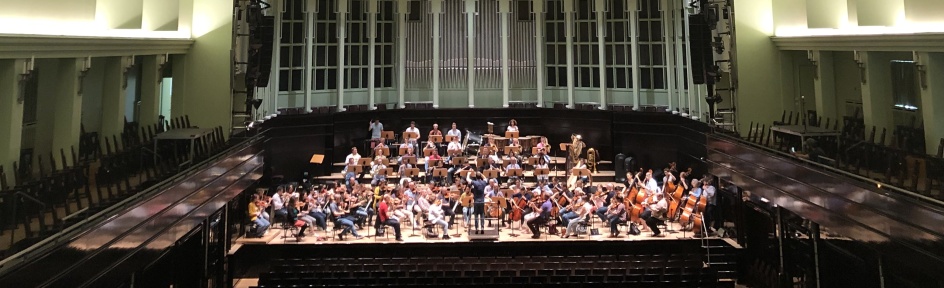 Ein Orchestra spielt in einem großen, leeren Konzertsaal. Der Saal