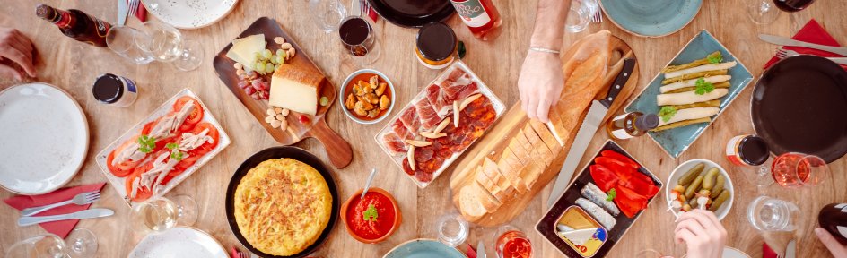 Ein gedeckter Tisch von oben, an dem sechs Menschen sitzen. Zu sehen sind verschiedene spanische Lebensmittel, wie Schinken, Käse und Wein. 