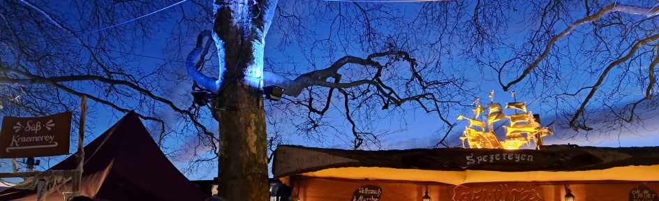 Buden beim historischen Markt auf dem Schlachte-Zauber an der Weserpromenade. Abendstimmung, die Bäume sind blau angestrahlt.
