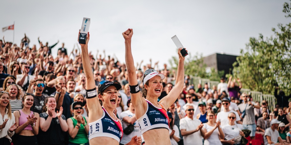 Two women cheer and hold up a trophy. Grandstand in the background.