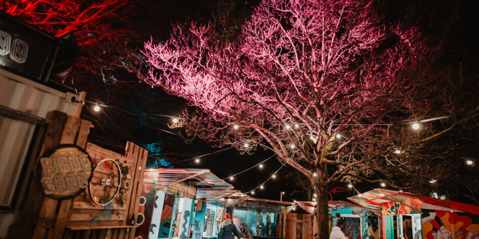Winter market stalls made of planks where people walk and stroll past and a festively illuminated tree. 