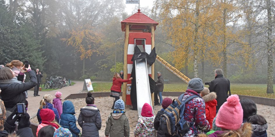Auf dem Bild ist ein Spielplatz in einem Park zu sehen. Senatorin Anja Stahmann enthüllt ein Spielgerät in Form eines Leuchtturms. Im Publikum sind viele Kinder zu sehen. Das Spielgerät ist mit Klettermöglichkeiten und Rutsche ausgestattet. Auf dem Dach befindet sich eine Fahne mit der Aufschrift "Platz der Kinderrechte".