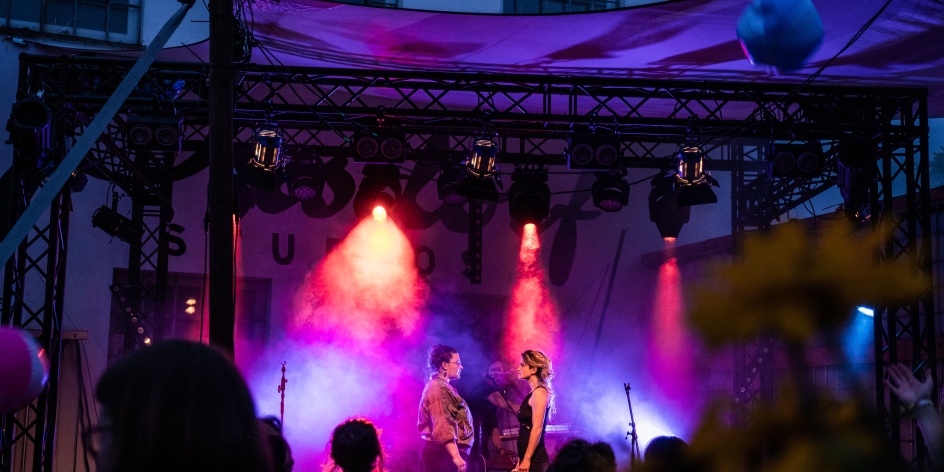 Zwei Personen stehen sich gegenüber auf einer Bühne, die in lila Licht und Nebel getaucht ist.