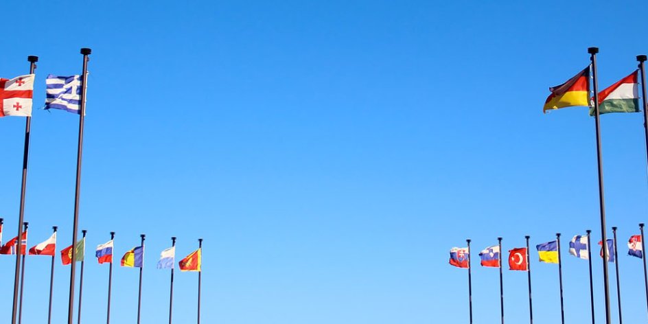 Unterschiedlichste Fahnen aus verschiedenen Ländern wehen im Wind vor blauem Himmel. Sie sind an Maste im Kreis arrangiert.