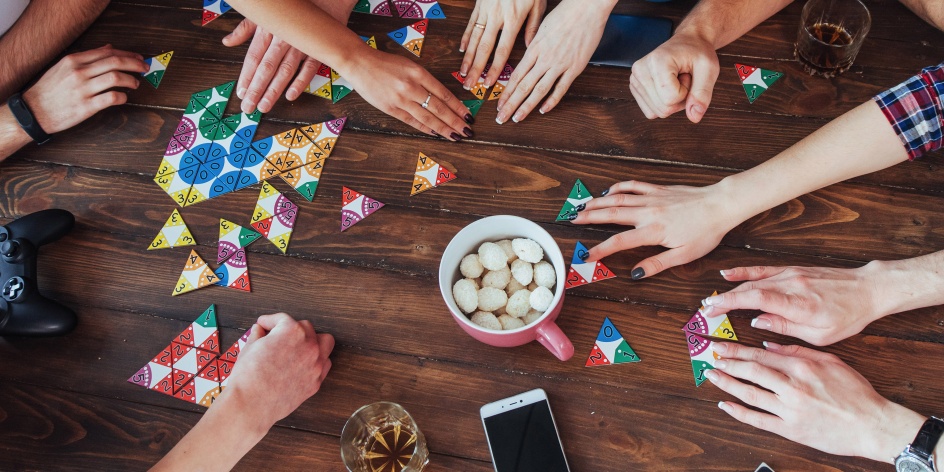 Das Foto zeigt die Hände von verschiedenen Menschen, die an einem Tisch ein Gesellschaftsspiel spielen. Die Aufnahme wurde von oben gemacht. 