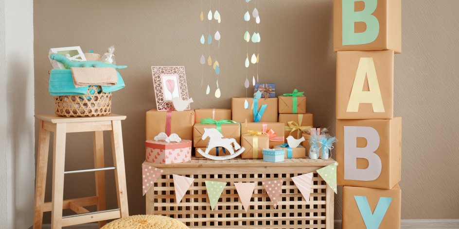 Eine Holzbox auf der eingepackte Geschenke liegen. Rechts von der Holzbox stehen übereinander gestapelte Boxen auf denen das Wort Baby steht. Auf der linken Seite steht ein Holzstuhl mit Handtüchern und einem Stoff-Dinosaurier drin.