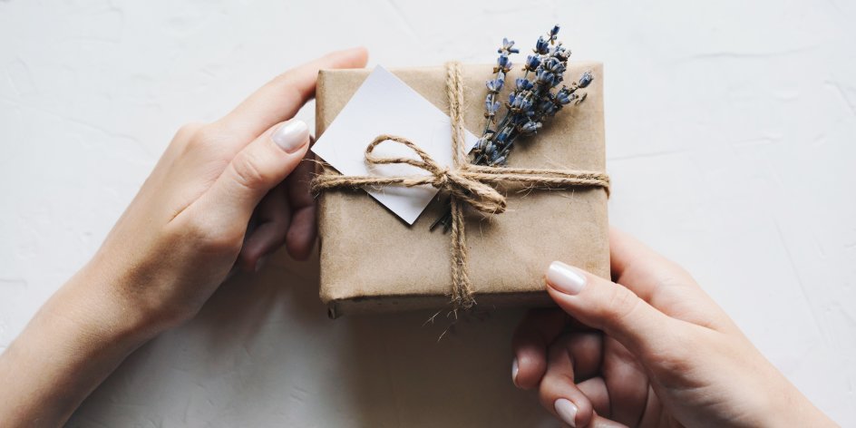 Auf weißem Hintergrund halten zwei Hände ein Geschenk, das in braunem Papier eingewickelt ist und mit einer Kordel, Lavendel und einer kleiner karte dekoriert wurde. 