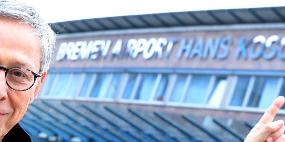 Ein Mann mit runder Brille und Krawatte zeigt auf den neuen Namen am Flughafenterminal