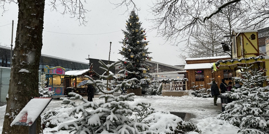 Auf einem Platz steht ein kleiner Weihnachtsmarkt mit einem Tannenbaum in der Mitte und es schneit.