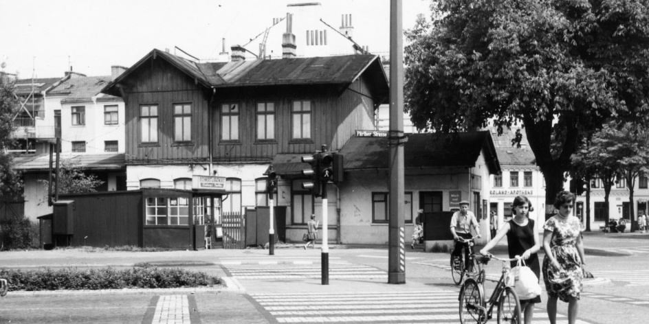 Zwei Frauen laufen über einen Zebrastreifen. Im Hintergrund ist auf der schwarz-weißen Aufnahme ein Bahnhof zu sehen. 