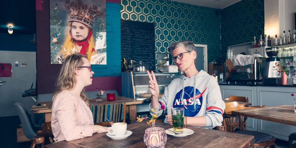 Zwei Frauen unterhalten sich in Gebärdensprache. Sie sitzen an einem Tisch in einem Café.
