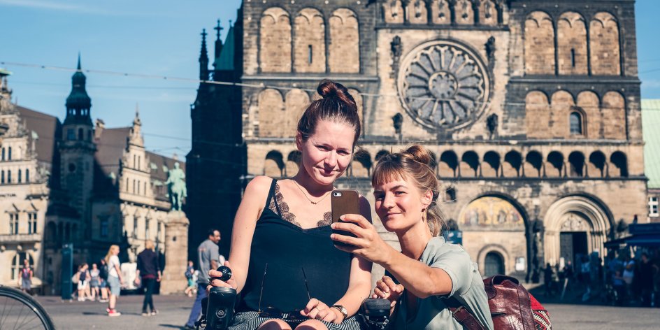 Zwei Frauen auf dem Bremer Marktplatz. Sie machen ein Selfie mit ihrem Smartphone, im Hintergrund ist der Bremer Dom zu sehen. Eine von den beiden Frauen sitzt im Rollstuhl.