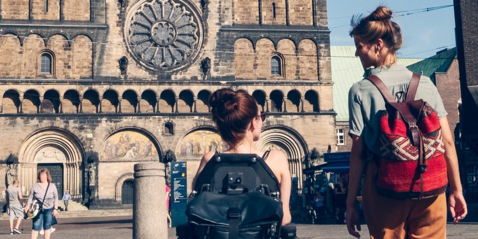 Zwei Frauen auf dem Bremer Marktplatz. Sie blicken auf den Dom und unterhalten sich. Eine von ihnen sitzt im Rollstuhl.