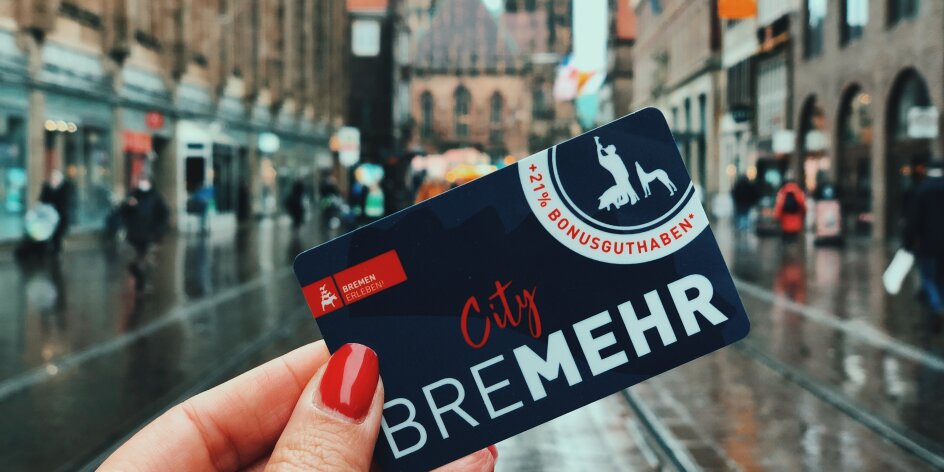 Eine Hand mit rot lackiereten Fingernägeln hält den "BreMEHR"-Gutschein vor der Kulisse der Bremer Innenstadt. 