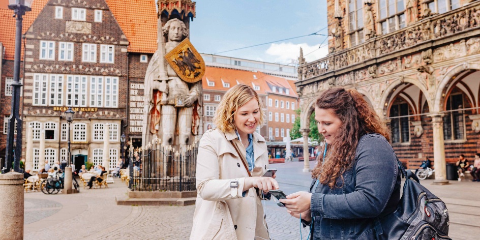 Two women look at the audio guide on Bremen's market square with headphones in their ears.