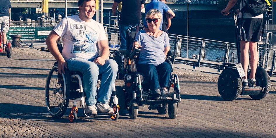Ein lächelnder Mann und eine Frau im Rollstuhl an der Schlachte. Außerdem sind Menschen auf Segways unterwegs.