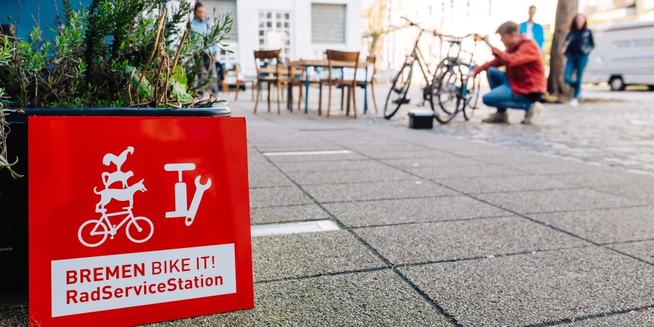 Logo Bremen BIKE IT! Rad Service Station. Im Hintergrund wird ein Fahrrad repariert.