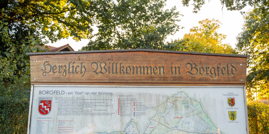 Willkommensschild am Ortseingang von Bremen-Borgfeld mit Karte und Informationen zum Stadtteil, umgeben von Bäumen im warmen Abendlicht.