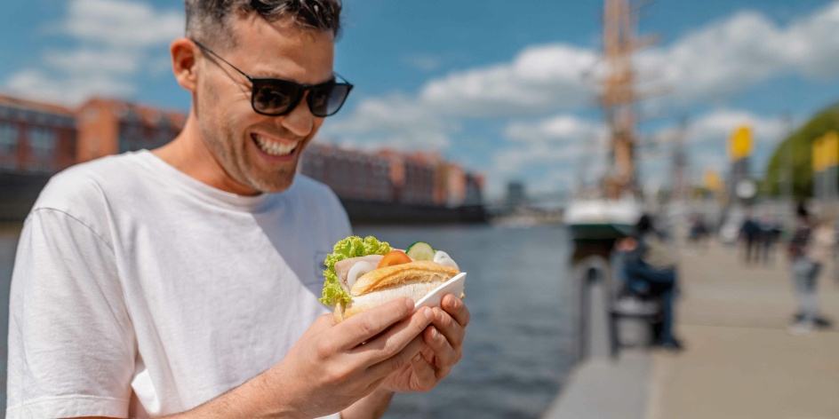 Ein Mann steht mit einem Fischbrötchen an der Schlachte. Es ist sonniges Wetter.