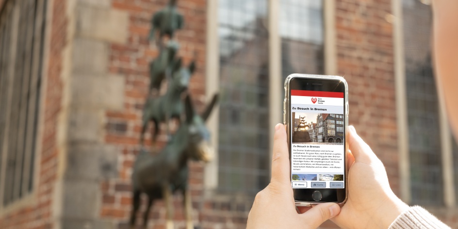 Jemand hält ein Smartphone vor den Stadtmusikanten. Auf dem Bildschirm sieht man die Startseite von "Dein Bremen Guide".