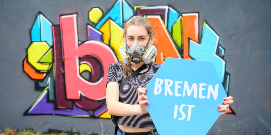 Eine Frau mit Atemmaske steht vor einem bunten Graffiti. In der Hand hält sie ein Schild mit "Bremen ist".