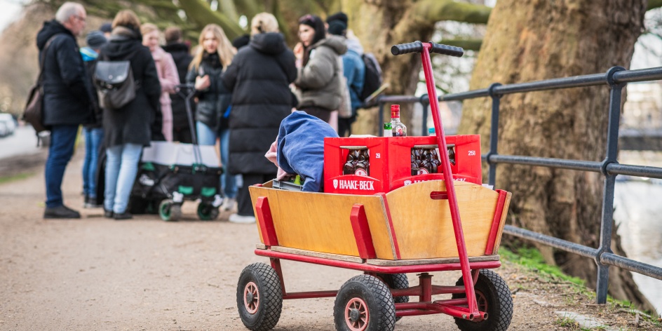 Ein Bollerwagen steht auf einem Weg. Im Hintergrund ist eine Gruppe zu sehen.