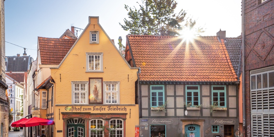 Zwei kleine historische Häuser. Über den Dächern strahlt die Sonne.