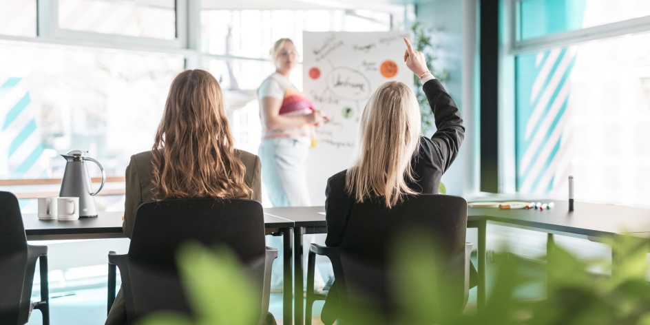 Eine Frau erklärt etwas an einem Flipchart, zwei weitere Frauen sitzen davor und eine davon meldet sich. 