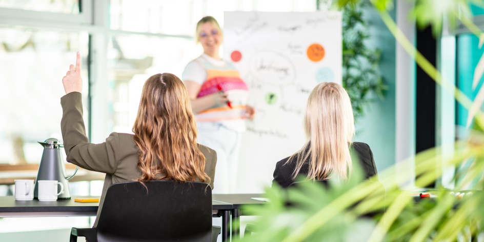 Eine Workshop-Situation zeigt zwei Teilnehmende von hinten, die dem Vortrag einer dritten folgen, die vor ihnen steht.