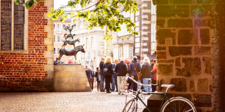 Die Stadtmusikanten Statue ist umringt von Menschen, im Vordergrund steht ein Fahrrad an die Wand gelehnt und Äste mit grünen Blättern hängen ins Bild herein.