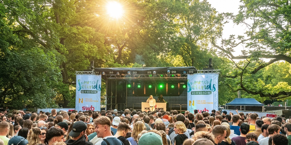 Eine Open-Air Bühne vor der ein großes Publikum steht. Hinter der Bühne sind große grüne Bäume und die Sonne scheint.