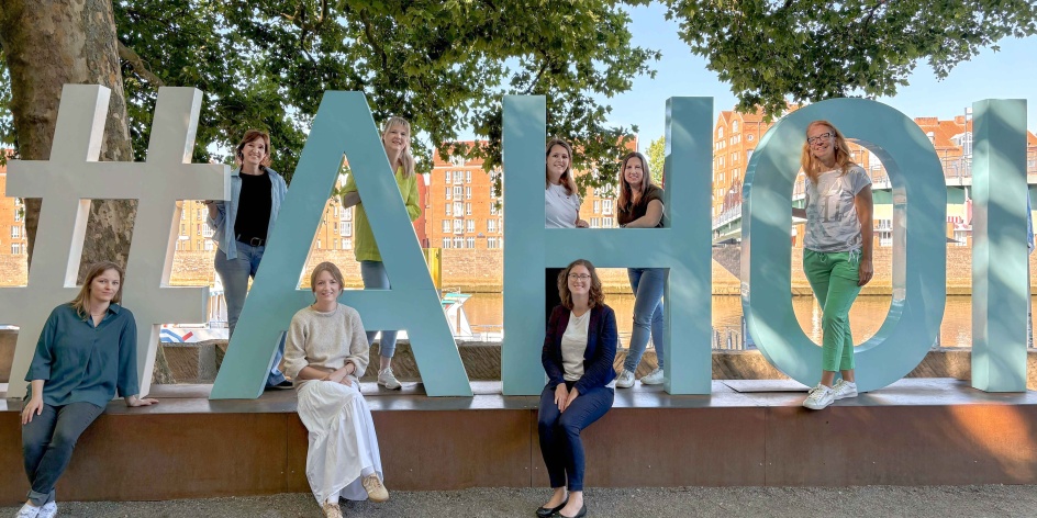 Acht Frauen stehen verteilt vor, hinter und zwischen großen Moin-Buchstaben und lächeln in die Kamera.