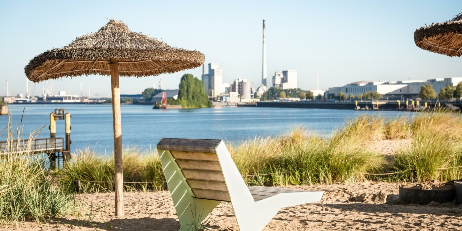 Der Blick auf den Waller Sand zeigt einen Liegestuhl und einen Sonnenschirm mit Palmenblättern an einem ausgedehnten Sandstrand vor der Kulisse des Bremer Getreidehafens bei Sonnenschein