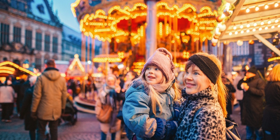 Eine Frau hat ein Mädchen auf dem Arm, sie steht vor einem Karussell auf dem Bremer Weihnachtsmarkt.
