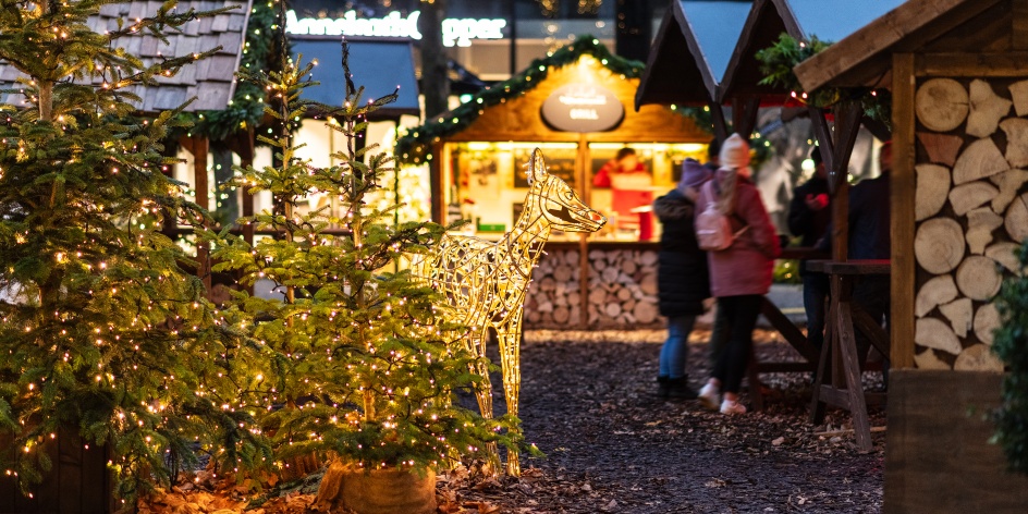 Tannenbäume und beleuchtete Buden auf einem Wintermarkt, Menschen stehen an einem überdachten Tisch. 