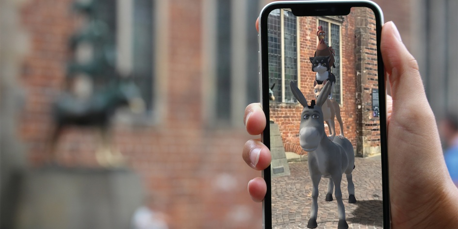 In the foreground, the Bremen Town Musicians can be seen in 3D through a smartphone. The statue of the Town Musicians can be seen in the background. 