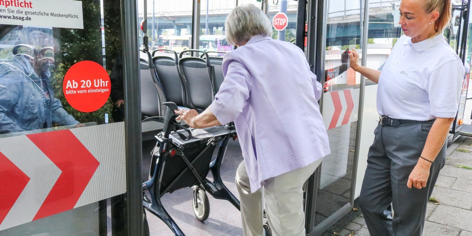 Eine ältere Dame mit Rollator steigt in einen Bus ein. Eine Mitarbeiterin der BSAG begleitet sie dabei.