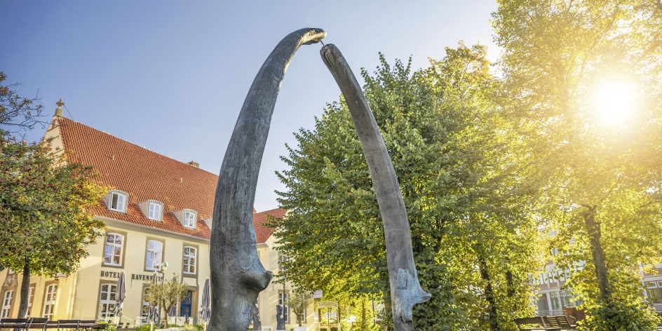 Ein kleiner Platz wird von einem Bogen geziert. Dahinter stehen grüne Bäume und ein Haus. Der Himmel ist blau und die Sonne scheint durch die Bäume.