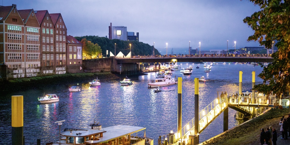 Auf dem Fluss "Weser" sind mehrere beleuchtete Schiffe zu sehen. Einige Menschen stehen auf einer Brücke und schauen den Schiffen zu. Es ist Abenddämmerung.