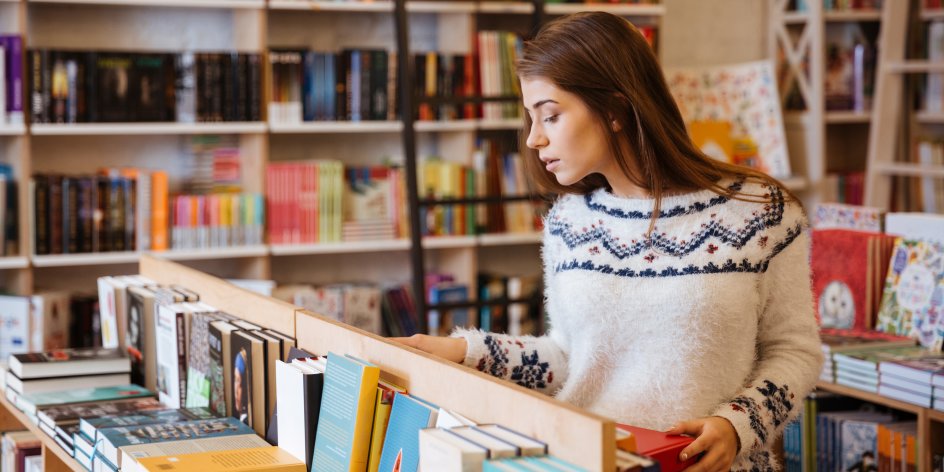 A young woman is standing in front of a table-high shelf in a bookshop, looking at the books. In the background are large bookshelves.
