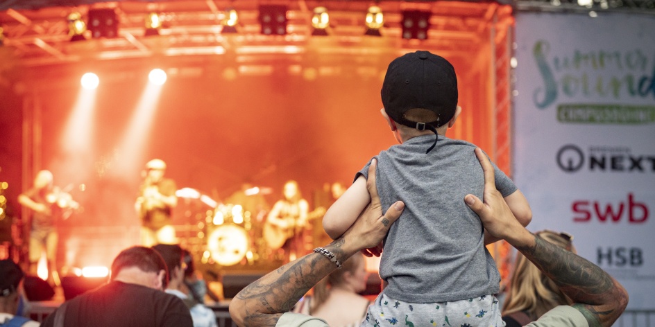 Ein Mann hat seinen Sohn auf den Schultern und schaut auf eine Bühne. Dort spielt eine Band. 