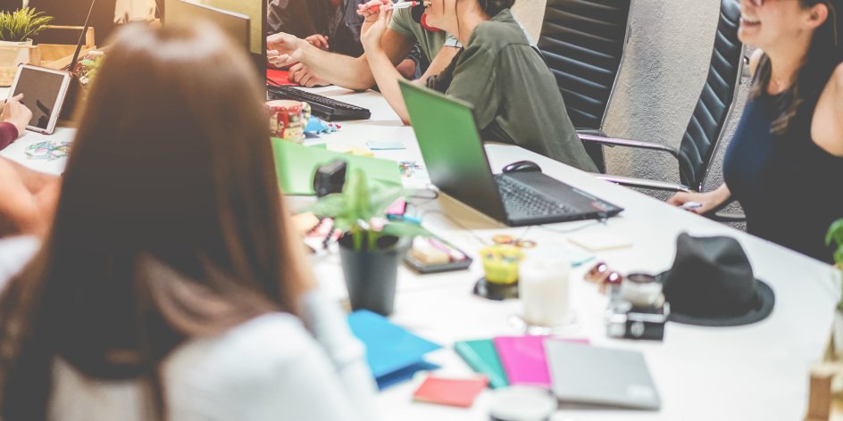 Junge, gut gelaunte Leute sitzen an einem großen Besprechungstisch und tauschen sich aus. Auf dem Tisch stehen Laptops und andere Arbeitsutensilien.