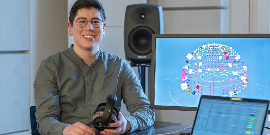 Ein Wissenschaftler schaut lächelnd in die Kamera. Auf der rechten Seite des Bildes befinden sich zwei Bildschirme, auf denen eine Grafik und eine Tabelle dargestellt wird. Hinter dem Wissenschaftler steht eine Lautsprecherbox.