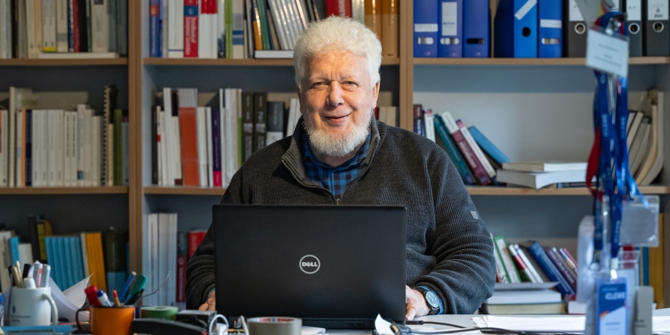 Klaus Boehnke sitzt an einem Schreibtisch und arbeitet an einem Laptop. Auf dem Tisch liegen Büromaterialien, hinter ihm befinden sich Regale mit Büchern und Ordnern.