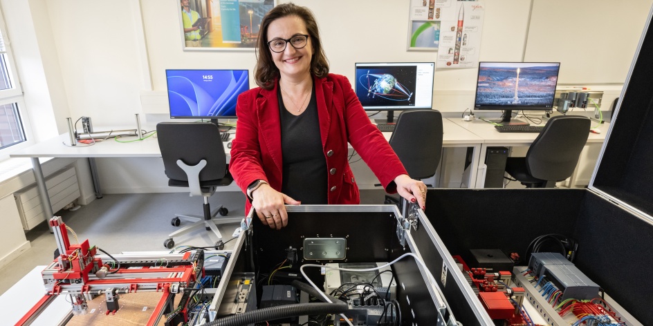 Die Wissenschaftlerin Jasminka Matevska steht in einem Arbeitsraum. Im Hintergrund stehen Bildschirme und vorne befindet sich eine Maschine. 