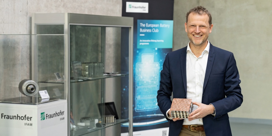 Ein Mann in einem dunkel blauen Sakko hält ein metallenes Exponat in der Hand. Neben ihm steht eine Glasvitrine mit weiteren Exponaten.  
