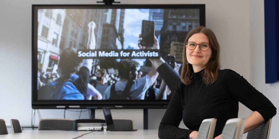 Eine Frau sitzt vor einem großen Monitor an einem Tisch. Darauf steht: Social Media for Activists