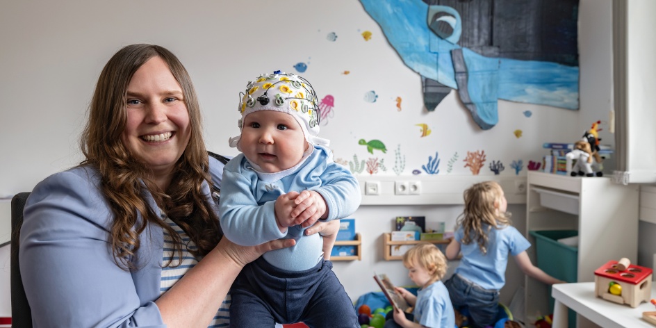 Eine Junge Frau hat ein Baby auf dem Arm. Im Hintergrund spielen zwei kleine Kinder.